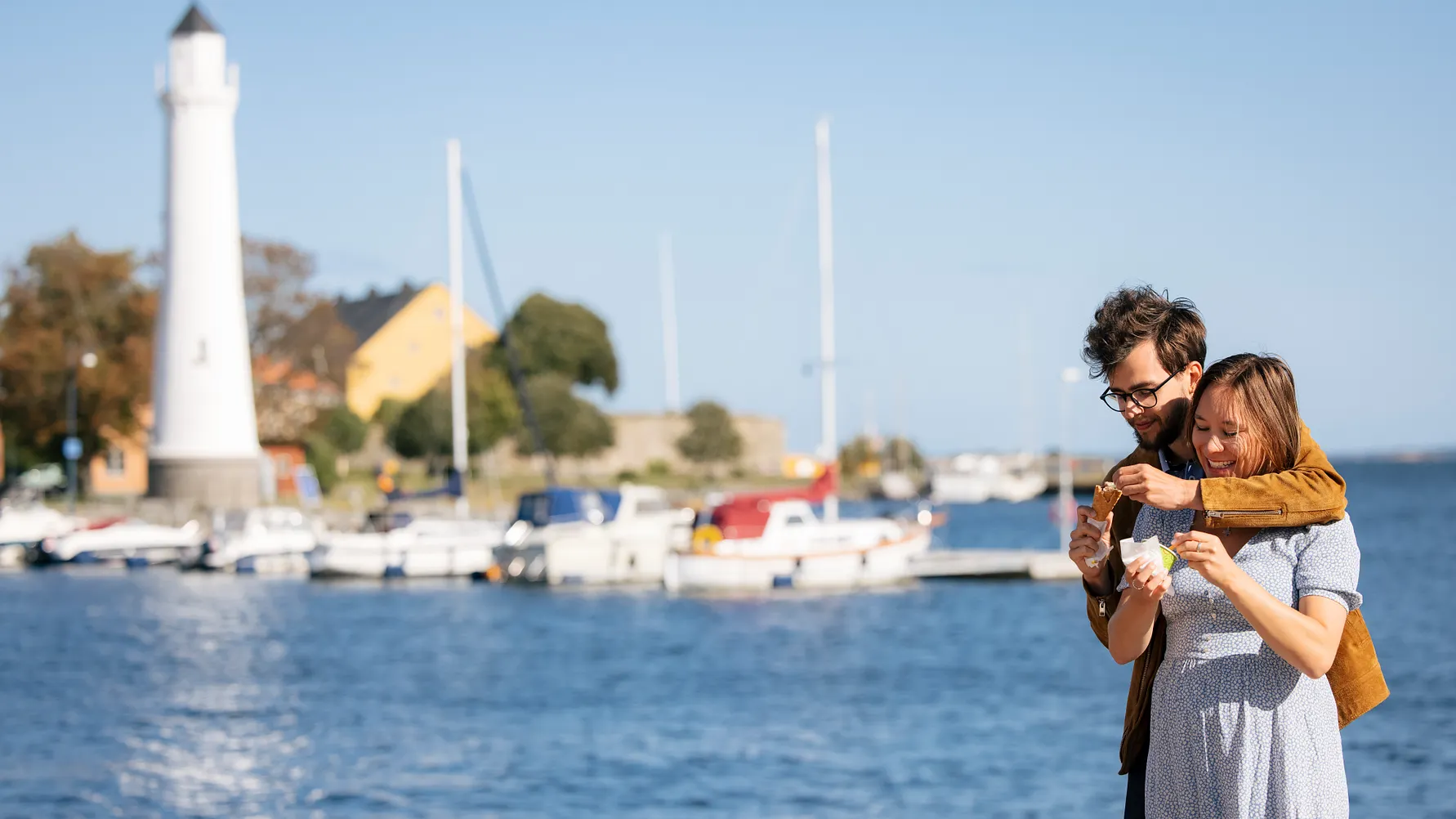 Bilden visar ett ungt par som äter glass vid havet. I bakgrunden syns båtar och en fyr.