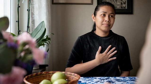 Bilden visar en kvinna vid ett köksbord. På bordet finns gröna äpplen i en korg och blommor. Kvinnan sitter med ryggen mot en vägg. 