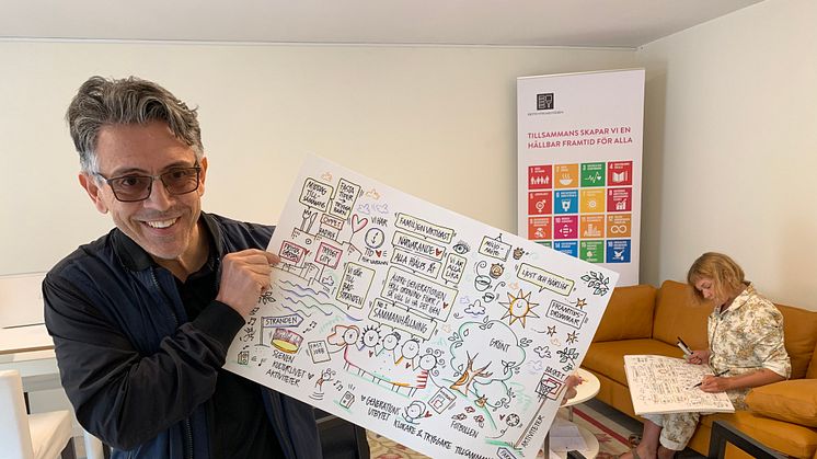 Alvaro Fuentes, hållbarhetsutvecklare på Botkyrkabyggen, med en av illustrationerna från Öppet hus i Fittja 24 maj. I bakgrunden illustratören Maja på Näset.