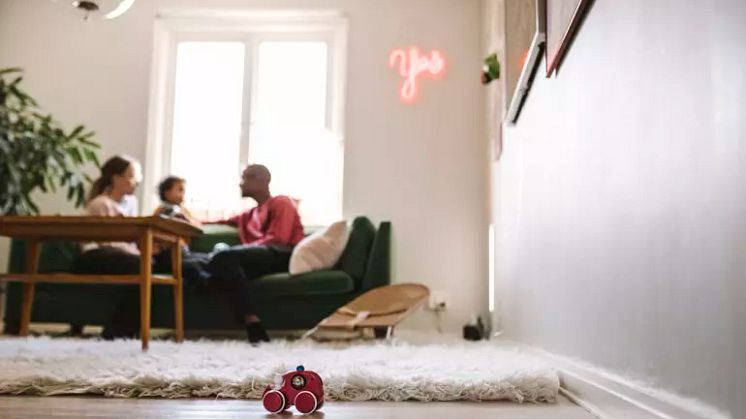 Mamma, barn och pappa sitter i en soffa i ett vardagsrum. Bilden är suddig och visar en leksaksbil i trä i förgrunden. 