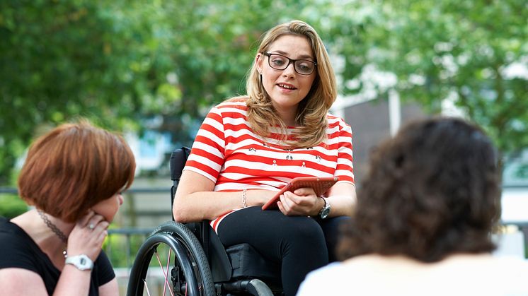 Ung kvinna i rullstol med mobil i handen talar med kompisar i en park.