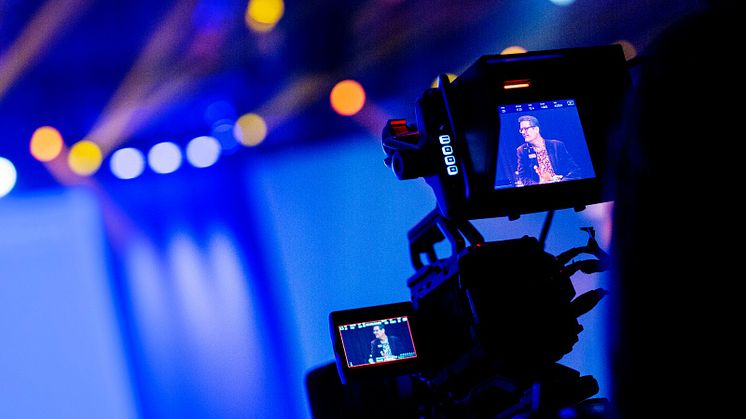 En videokamera står riktad mot en blåaktig scen med gula och vita lampor, i skärmen syns en man som ler. Foto: Apelöga/MTM