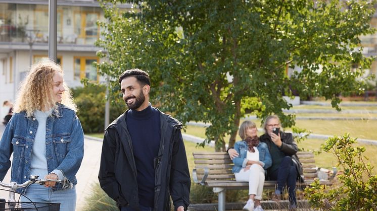 Syntolkning: Ett par på promenad, äldre par på en parkbänk i bakgrunden