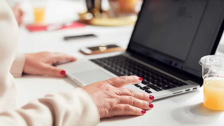 En persons händer med rött nagellack. Personen sitter vid ett bord med en laptop framför sig.