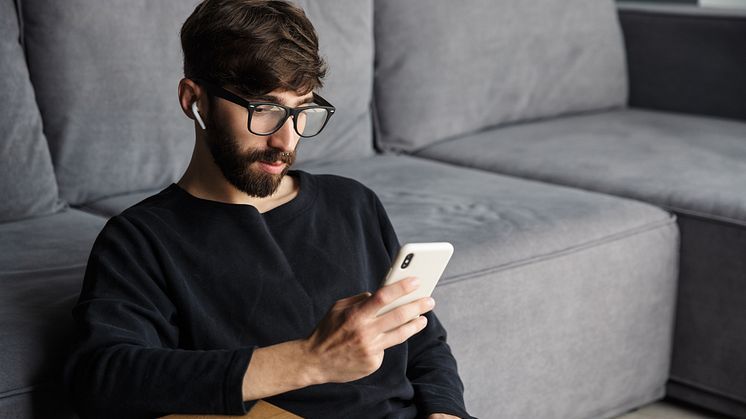 Syntolkning: Man med glasögon i hemmiljö tittar på en mobiltelefon
