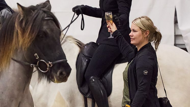 Tävlingsledaren för Agria Icelandic Power, Rebecca Jónasson, räcker en av de tävlande i Champagnetölten det glas som hon snart ska göra sitt bästa för att inte spilla ut. Samtidigt håller hon en öga på vad som pågår på banan.