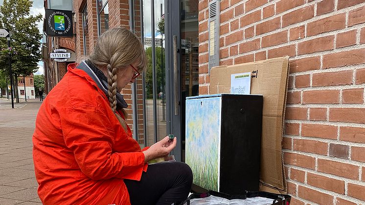 Konstnären Ann-Christine Jönsson sitter på plats på Mårtensgatan i Kävlinge och målar vackra grässtrån på ett elskåp. Fotograf: Monika Carnehult