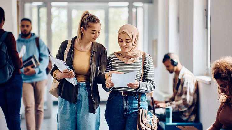 Happy Muslim student and her friend read their exam results while walking through university hallway (2)