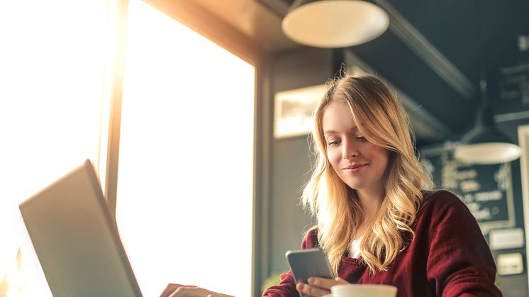 Ung kvinne på café som jobber på både mobil og laptop