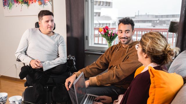 Personer sitter tillsammans och pratar. En person skriver på laptop. De är glada. En person är rullstolsburen. 