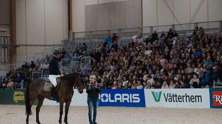 Kyra Kyrklund tillsammans med Peder Fredricson och Raadbjergs’s Bahia under clinicen på Jönköping Horse Show. Med hjälp av sin berömda fjäder illustrerar Kyra hästens rörelse vid ridning. 