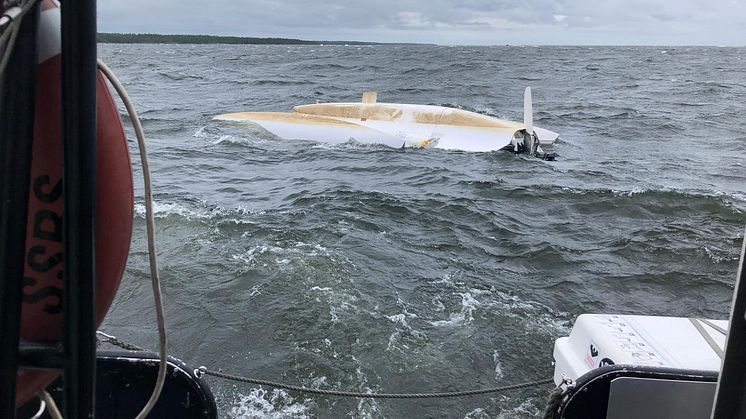 På den uppochnedvända båten, i nätet mellan skrov och ponton, sitter två kraftigt nedkylda personer. De har skickat upp en nödraket i hopp om hjälp.