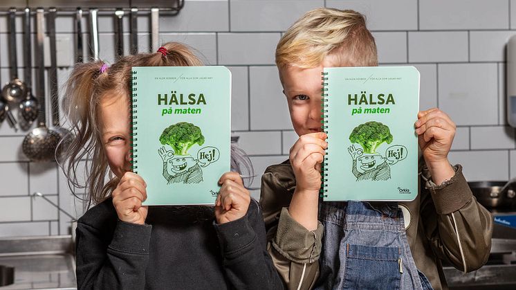 Inez och Max håller upp vars ett exemplar av kokboken "Hälsa på maten", framtagen för barn och pedagoger.