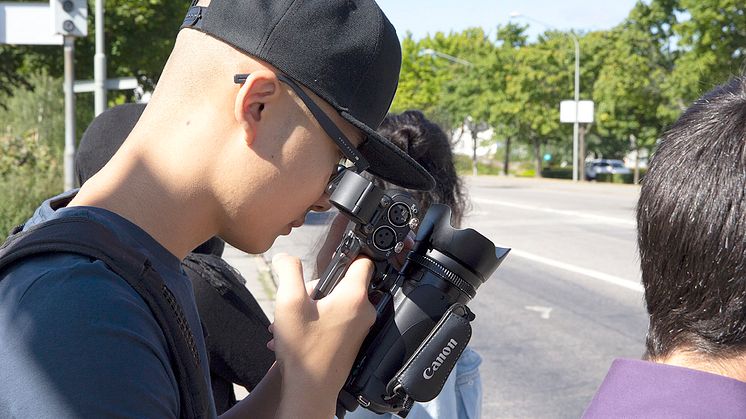 Syntolkning: Ungdomar utomhus vid en väg, en håller i och tittar in i en filmkamera. Foto: Mikael Ström