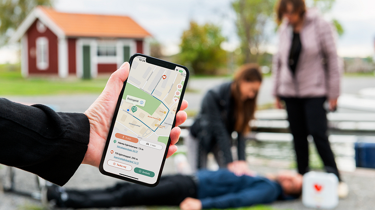 Person håller i telefon med appen Sms-livräddare öppen medan en kvinna utför Hjärt-lungräddning i bakgrunden 