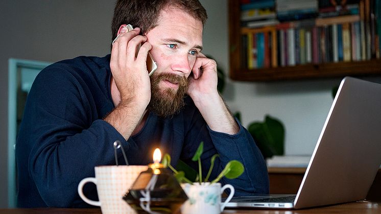 En man med skägg sitter vid ett bord i hemmiljö framför en dator och pratar i telefon. På bordet står ett ljus och en tekopp.