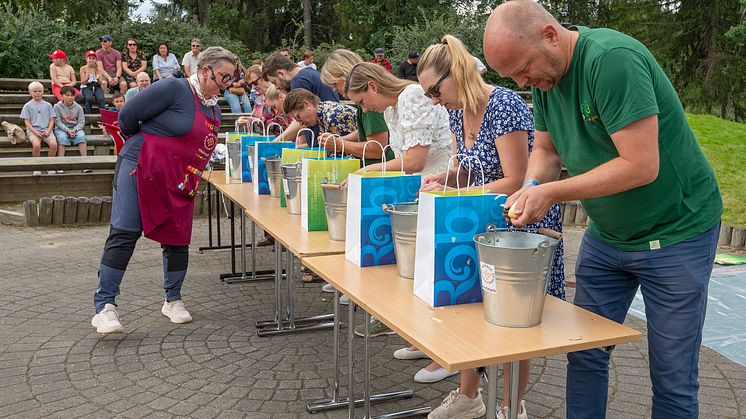 En rekke med mennesker som står ved et langbord og skreller poteter i hver sin bøtte.