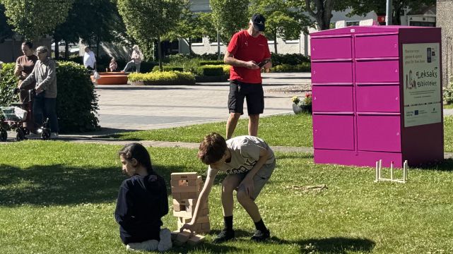 Barn leker i gräset med ett super-jenga spel framför en rosa Pifflbox från Leksaksbiblioteket. 