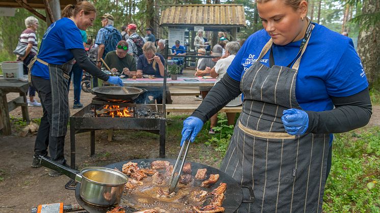 En ung kvinne med forkle som tilbereder flesk i en stor panne utendørs.