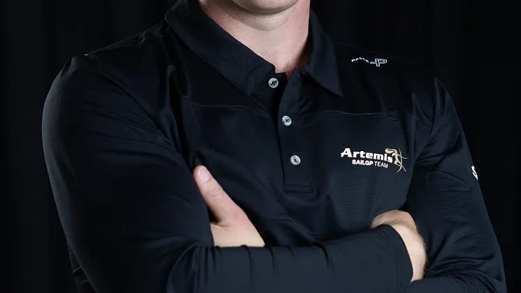 Leende man med armarna i kors, klädd i en svart tröja med logotypen Artemis SailGP Team, fotograferad mot mörk bakgrund.