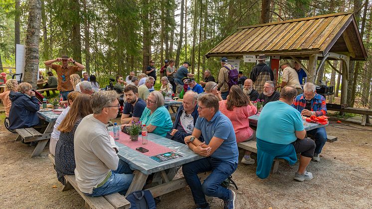 Mange folk som sitter og koser seg rund bord ved et serveringssted omgitt av skog.