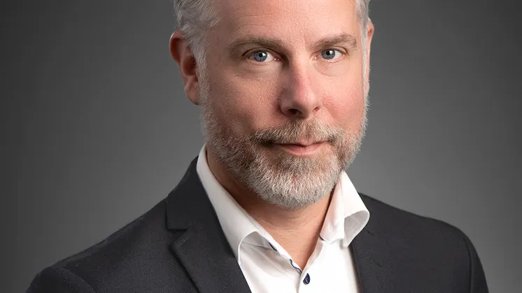Christian har kort ljust hår, skägg, blåa ögon, vit skjorta och grå kavaj.