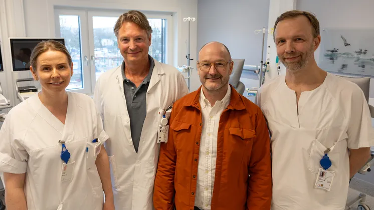 En kvinnelig sykepleier, mannlig lege, pasient (voksen mann med oransje jakke og briller) og mannlig kirurg står i et behandlingsrom på sykehus.