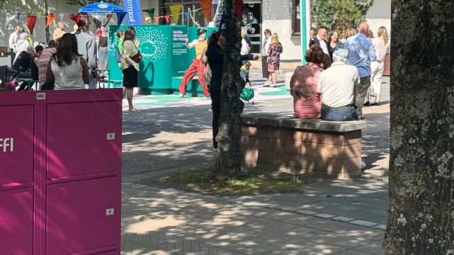  En rosa Piffl-aktivitetsbox står vid en park där människor samlas. Träd kastar skuggor över boxen, och i bakgrunden syns bän