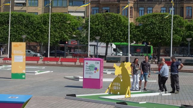 Pifflbox på Sundstorget i Helsingborg med människor som spelar minigolf runt omkring, solig dag och grönskande träd i bakgrun