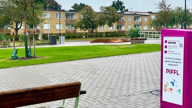 Rosa Pifflbox placerad vid torget fjärilen i Nödinge som ligger i Ale kommun. Med bänkar, träd och bostadshus i bakgrunden.