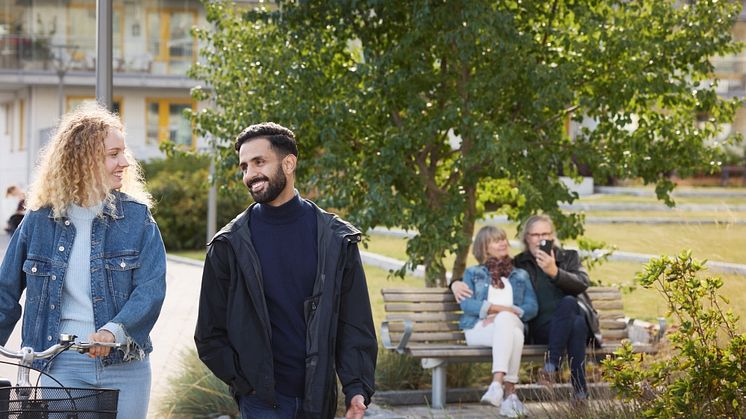 Syntolkning: Ett par på promenad, äldre par på en parkbänk i bakgrunden
