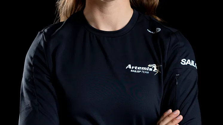 Kvinna med armarna i kors, klädd i en svart tröja med logotypen Artemis SailGP Team, fotograferad mot mörk bakgrund.