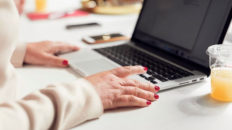 En persons händer med rött nagellack. Personen sitter vid ett bord med en laptop framför sig.