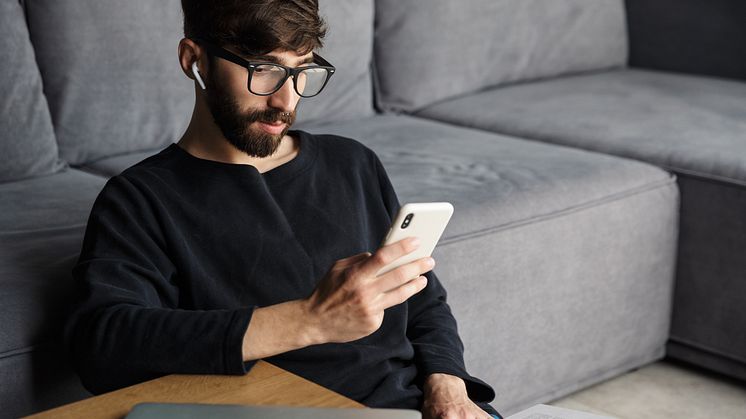 Syntolkning: Man med glasögon i hemmiljö tittar på en mobiltelefon