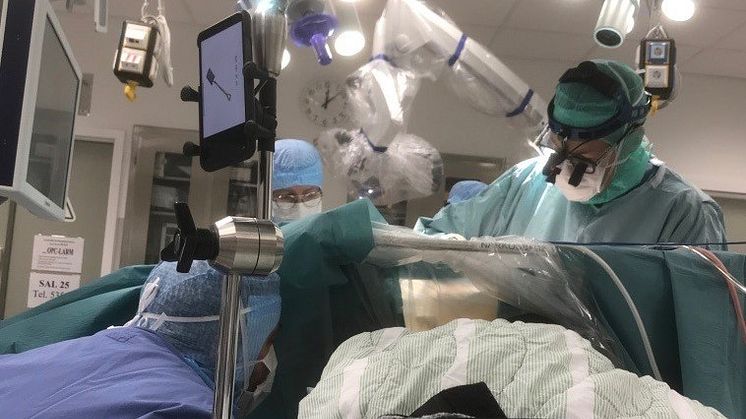 Neuropsykolog Mattias Stålnacke som samtalar med en patient medan denne opereras av överläkare Rickard L Sjöberg och specialistläkare Sajedha Mahmood och operationssjuksköterska Victoria Sagehem. I förgrunden syns testplatta.