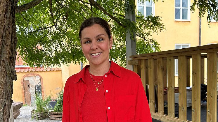 Lina Ejlertsson står under ett lövträd och ser in i kameran. Hon har på sig en röd tröja, röd skjorta och vita byxor.