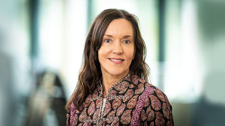 Susanna Toivanen tittar in i kameran. Hon har långt, brunt hår och en lilamönstrad skjorta på sig. Bakgrunden är suddig.
