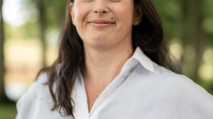 Leende kvinna i vit blus som står i en grön allé.