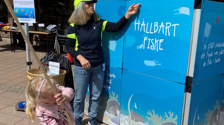En flicka fiskar i en blå fiskdamm med texten Hållbart fiske. En kvinna i gul keps hjälper till med en hov.