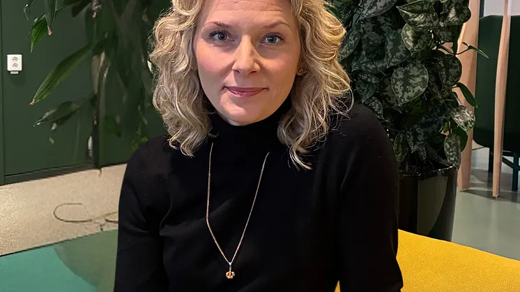 Anna Williamsson har ljust, utsläppt hår, svart tröja och ett halsband hon sitter i en gul soffa framför en stor grön växt. 