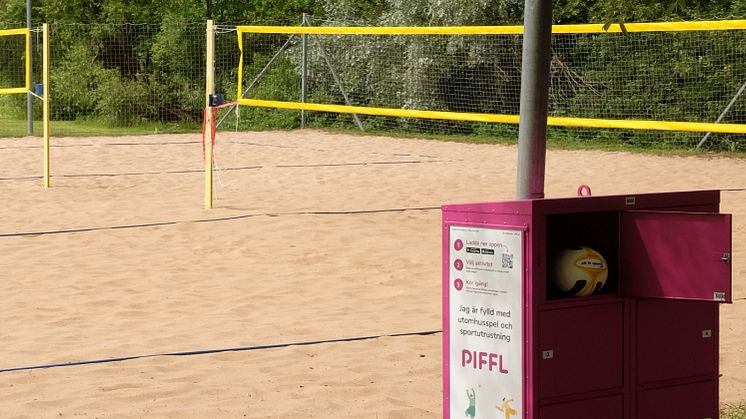 Pifflbox vid beachvolleyplan med nät och boll, omgiven av grönska i ett somrigt landskap i Linköping.  En volleyboll syns.