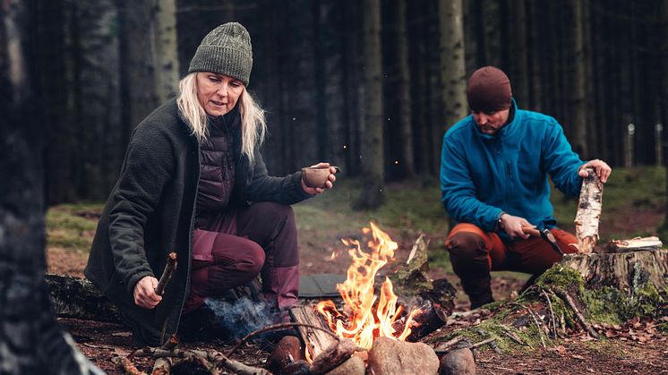 Kvinna och man i Pinewood AW25-kollektionen vid lägereld i skogen – hon dricker ur kåsa, han hugger ved.