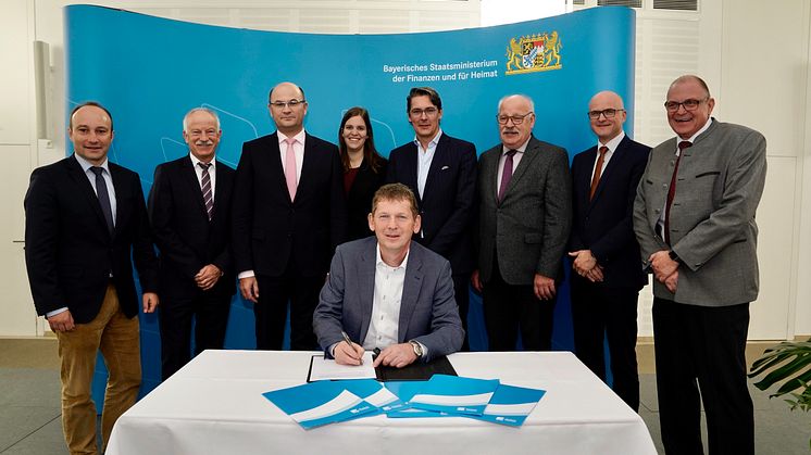 Michael Dümig von der Kommunalen Allianz Westspessart unterschreibt die Kooperationsvereinbarung im Beisein von Finanz- und Heimatminister Füracker (3. v.l.), Deutsche Glasfaser CEO Uwe Nickl (5. v.l.) und weiteren Vertretern der Allianz und DG.