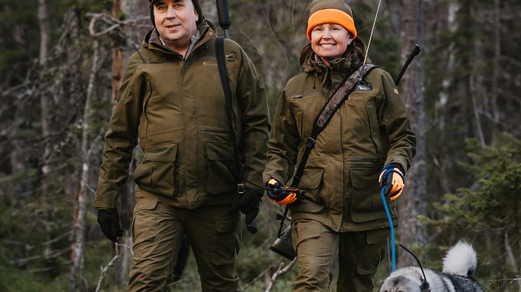 Två jägare i gröna jaktkläder från Pinewood går genom skogen med en älghund i koppel.