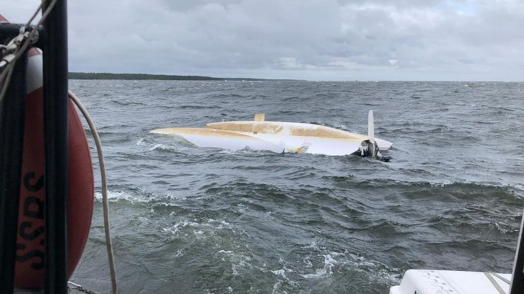 På den uppochnedvända båten, i nätet mellan skrov och ponton, sitter två kraftigt nedkylda personer. De har skickat upp en nödraket i hopp om hjälp.