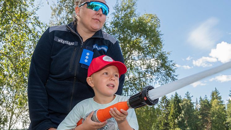 En kvinnelig offiser fra Sivilforsvaret hjelper en liten gutt å spyle med brannslange.