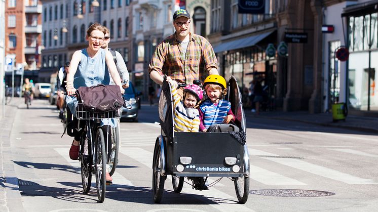 Cyklande man och kvinna i stadsmiljö där mannen har två barn i en cykelvagn.