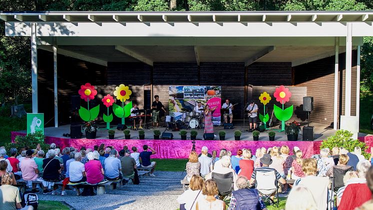 På den dekorerade scenen spelar artisterna musik i den grönskande parken. Framför scenen sitter publiken. Foto.
