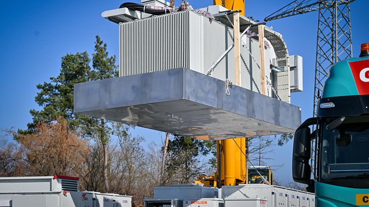 Anlieferung eines Transformators auf das Gelände des Wasserkraftwerks in Gersthofen. Im Hintergrund befinden sich die bereits aufgebauten Module des Batteriespeichers, der auf eine Speicherkapazität von rund 12 Megawattstunden ausgelegt ist.