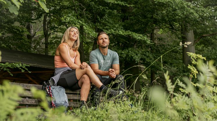 zwei Personen sitzen auf einer Holzbank inmitten Wald und Grün